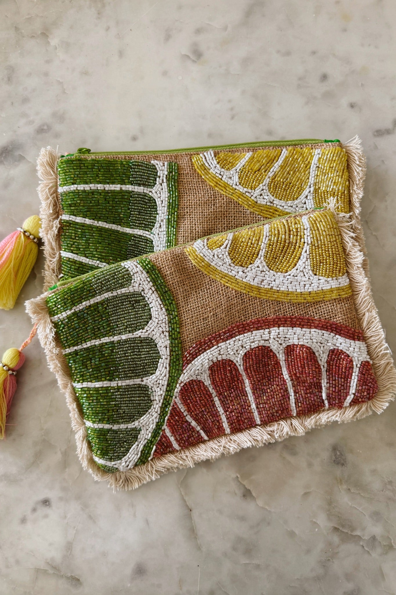 A vibrant Summer Citrus Jute Clutch by ANTIK KRAFT featuring a hand-beaded lemon slices design, fringes, and a zip closure. The clutch includes a fringe tassel detail and is lined for added style. It measures approximately 9.5” x 6.5” and is made from a blend of jute and cotton.
