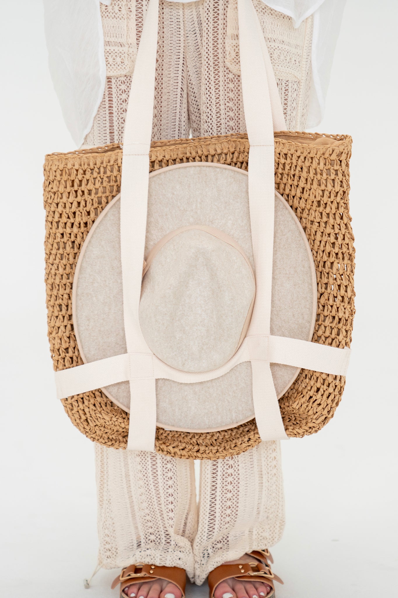 A tan raffia bag by Larrea Cove, known as the Ironwood Bag, featuring built-in hat holder straps. The bag has a spacious design suitable for beach vacations and summer outings. Its dimensions are 16 inches long, 17 inches high, and 1 inch wide.