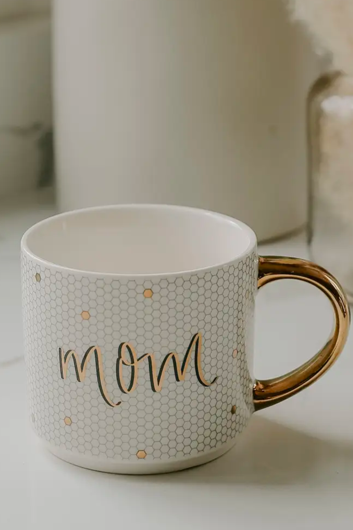 A stylish white mug featuring a honeycomb design and a gold handle, labeled "Mom" in elegant lettering. The mug holds 17oz and has a durable Fine Bone China construction. It is designed with a pattern on both sides and is suitable for both microwave and dishwasher use.