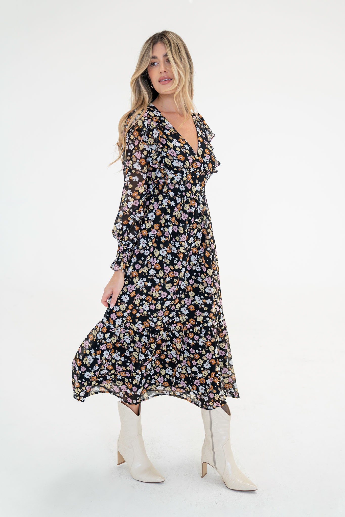 A model wearing the Pansies Floral Midi Dress by Larrea Cove, featuring a floral pattern, romantic ruffle shoulders, and a flattering V-neckline. The dress has a smocked side waist, an invisible back zipper, and is made of lightweight chiffon. The model is also wearing stylish sunglasses.