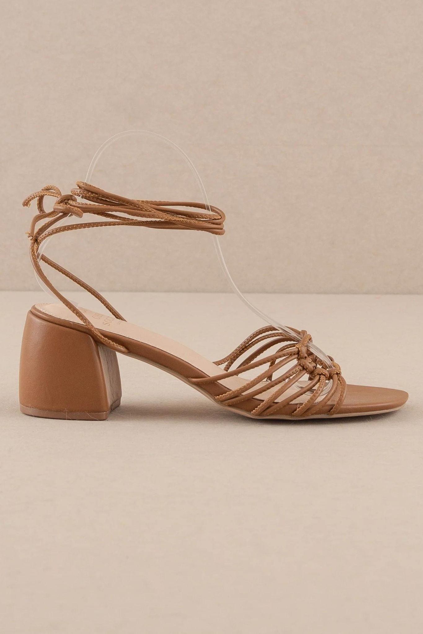 The Celia Brown Knotted Lace Up Heel by Oasis Society features a stylish brown leather design with knotted detailing and a lace-up front. These heels are perfect for summer occasions, showcasing a chic and trendy look. Accompanied by fashionable sunglasses, the ensemble represents the latest summer styles for 2024.