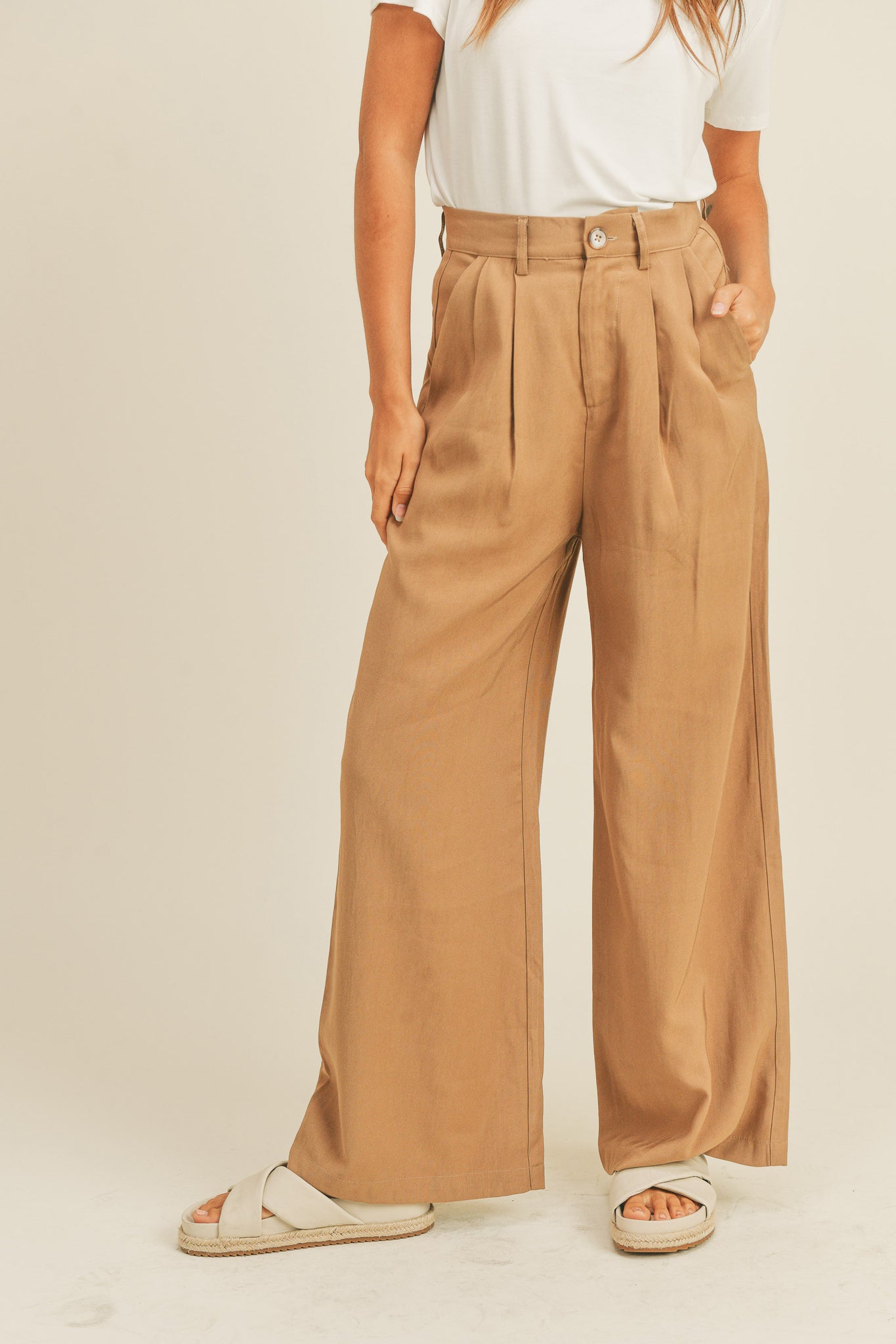 Camel-colored Arid Trousers by Larrea Cove featuring a wide leg, high waist, zip fly with button closure, and pleats at the top, displayed on a mannequin. The model is accessorized with sunglasses.