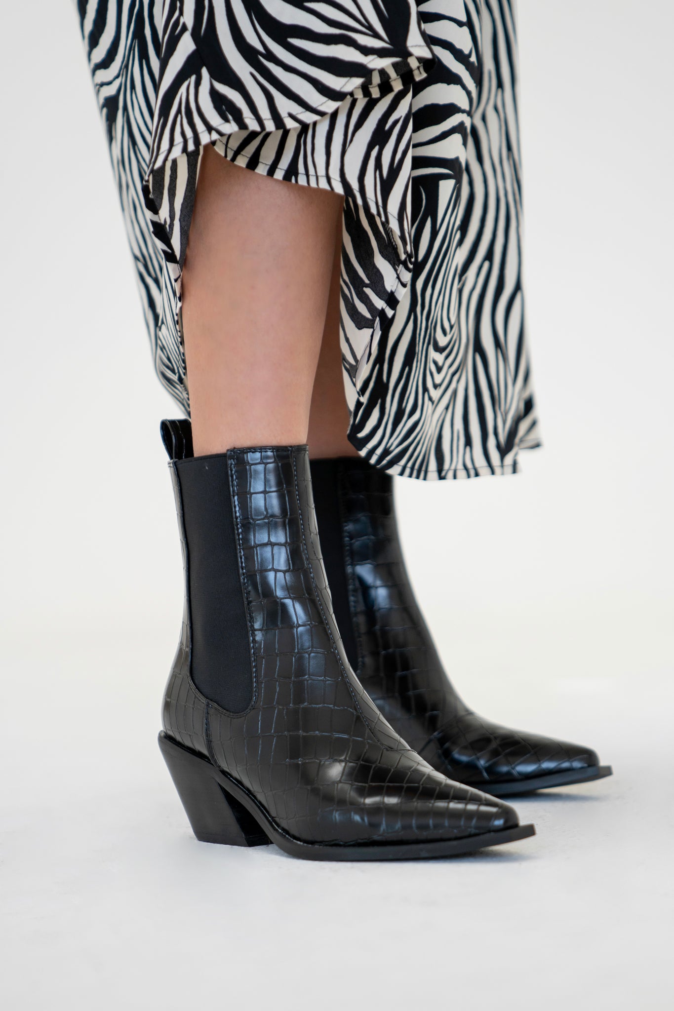 A pair of chic black ankle booties by Billini, featuring a mid stacked block heel, a squared off pointed toe, and a high cut upper. The boots have large elasticated side gussets and a back pull tab, showcasing a western-inspired design. The synthetic material gives a sleek appearance, ideal for transitioning from day to night looks.