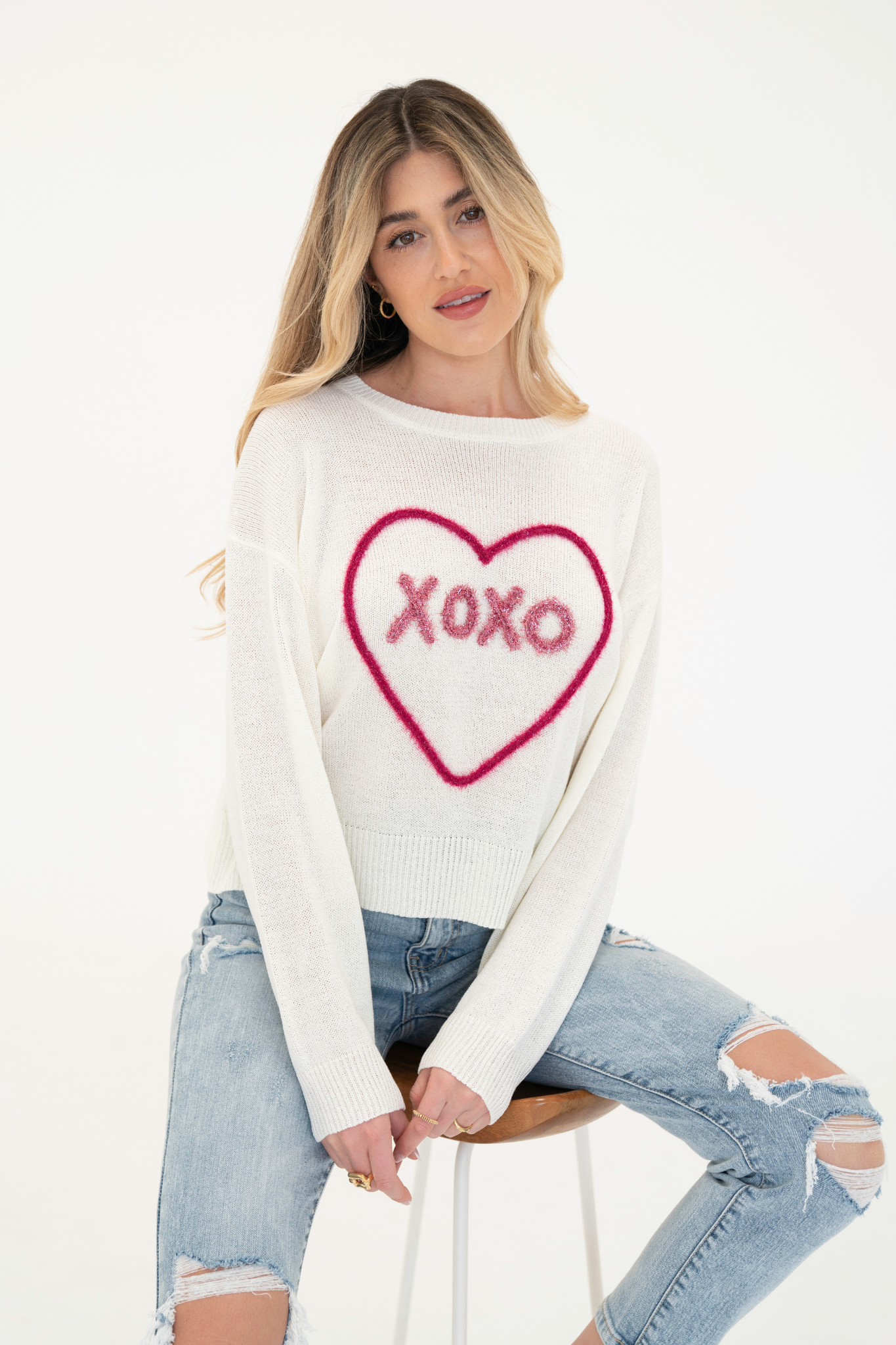 A model wearing the XOXO Sweater Top by Larrea Cove, featuring a lightweight, cozy design perfect for Valentine's Day. The sweater has a soft texture and a playful "XOXO" motif, embodying a romantic spirit. The model is 5'8" and wearing a size Small, with a casual style complemented by sunglasses.
