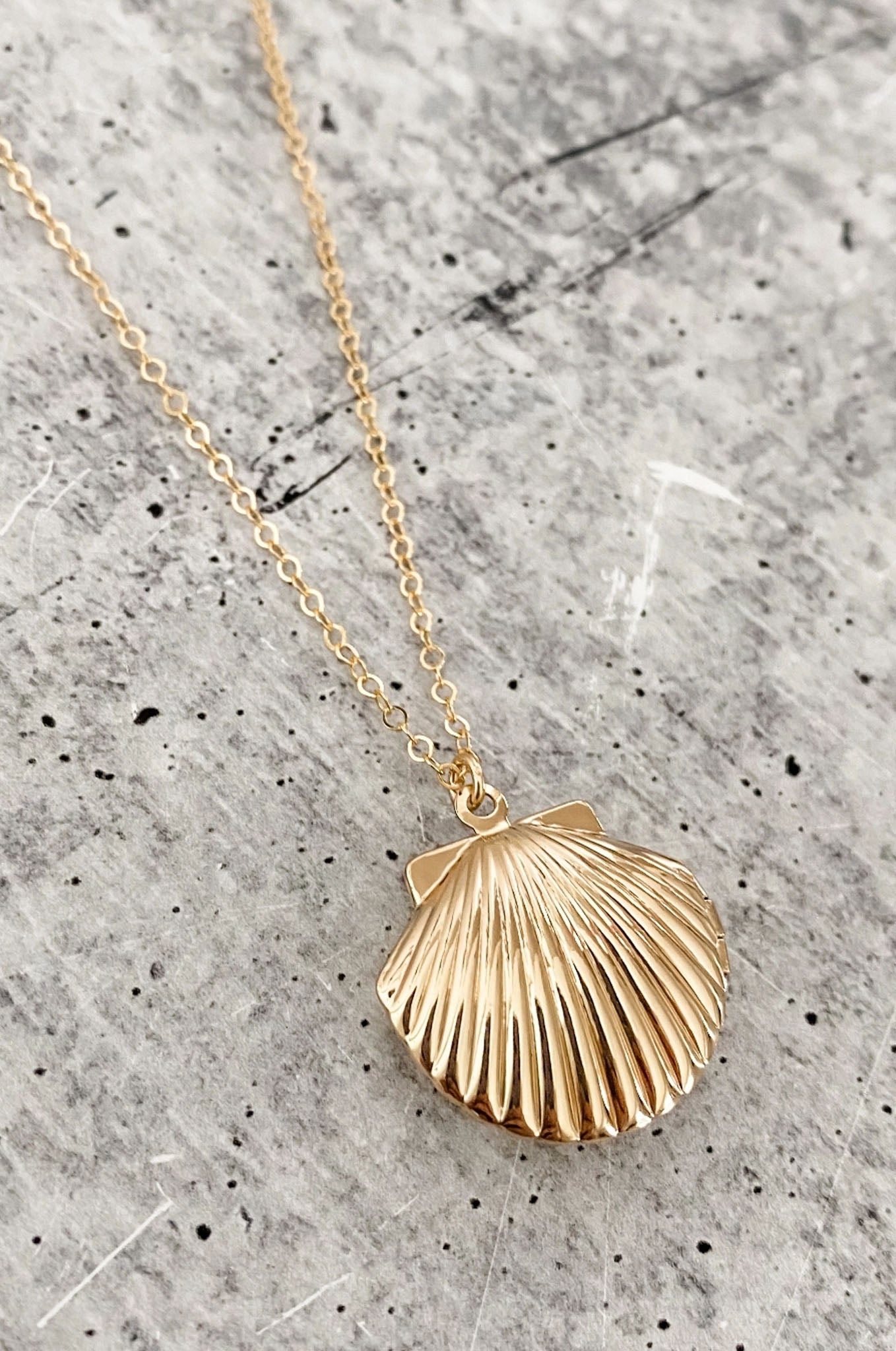 A vintage-style gold-plated scallop seashell locket necklace by Larrea Cove. The locket is approximately one inch wide and high, hanging from a shiny 14k gold-filled 18-inch cable link chain. The necklace has a delicate and sparkly appearance, showcasing a handmade design. Ideal for adding a classic touch to any outfit.