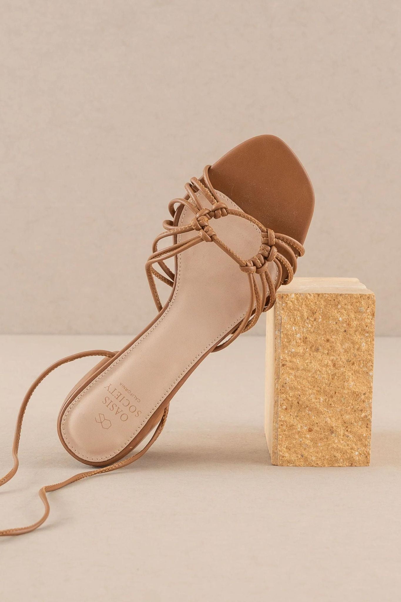 Image of the Celia Brown Knotted Lace Up Heel by Oasis Society. The shoe features a stylish brown hue with a knotted design and lace-up closure, perfect for summer. Accompanied by a pair of trendy sunglasses. Tags include Shoes, Summer 2024, and new product release for July 2024.